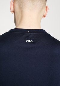 Ναυτικό μπλε αθλητικό πουκάμισο με υφή υφάσματος, διαθέτει ασημένιο λογότυπο στον πίσω λαιμό και μια μικρή μεταλλική λεπτομέρεια από πάνω.