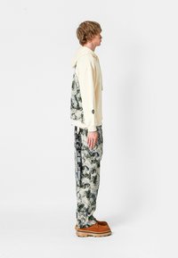 Sudadera cremosa clara con un respaldo texturizado y pantalones de camuflaje en tonos terrenales, con detalles en negro y un ajuste relajado. Zapatillas marrones completan el conjunto.