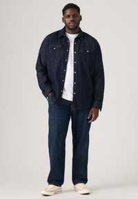 Mann in dunkelblauem Denimhemd, weißem T-Shirt, blauen Jeans und weißen Sneakers, der mit einer Hand in der Tasche vor einem einfarbigen Hintergrund steht.