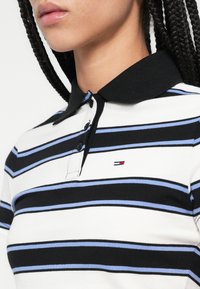 Csíkos pólóing fekete, fehér és világoskék csíkokkal, fekete gallérral és háromgombos patenttal, a mellrészen logóval.