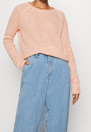 Donna che indossa un maglione lavorato a maglia rosa chiaro con maniche lunghe infilato nei jeans larghi a vita alta azzurri.