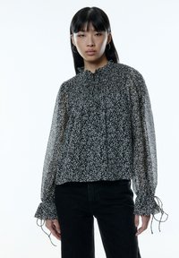 Zwarte bloemen sheer blouse met een verzamelde halslijn, lange mouwen en ruffled manchetten. Heeft een losse pasvorm en delicate witte patronen.