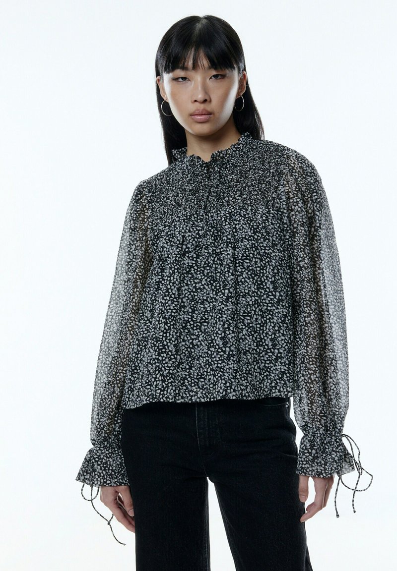 Zwarte bloemen sheer blouse met een verzamelde halslijn, lange mouwen en ruffled manchetten. Heeft een losse pasvorm en delicate witte patronen.