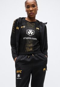 Tenue de sport noire composée d'un t-shirt à manches courtes avec accents dorés et d'un pantalon. Le t-shirt présente un design graphique et un logo ; le pantalon possède une ceinture élastique.