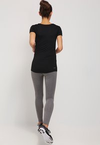 Femme de dos portant un T-shirt noir ajusté, des leggings gris et des chaussures de sport noir et blanc sur fond uni.