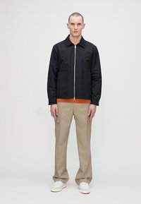 Sort zip-up jakke med to frontlommer, orange skjorte under, beige cargo-bukser og hvide sneakers. Glat stof og moderne pasform.