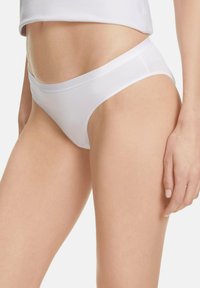 Kobieta w prostych białych bawełnianych majtkach w stylu bikini oraz białym topie na ramiączkach, pokazana od połowy torsu do górnej części ud.