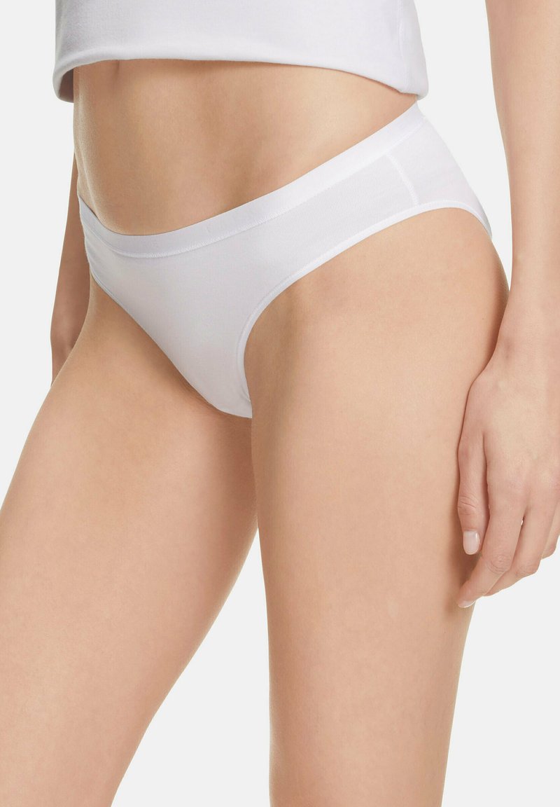 Kobieta w prostych białych bawełnianych majtkach w stylu bikini oraz białym topie na ramiączkach, pokazana od połowy torsu do górnej części ud.