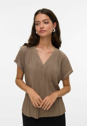 Femme aux longs cheveux noirs portant des boucles d'oreilles en argent et un blouse brun à col en V plissée à manches courtes, debout les mains jointes.