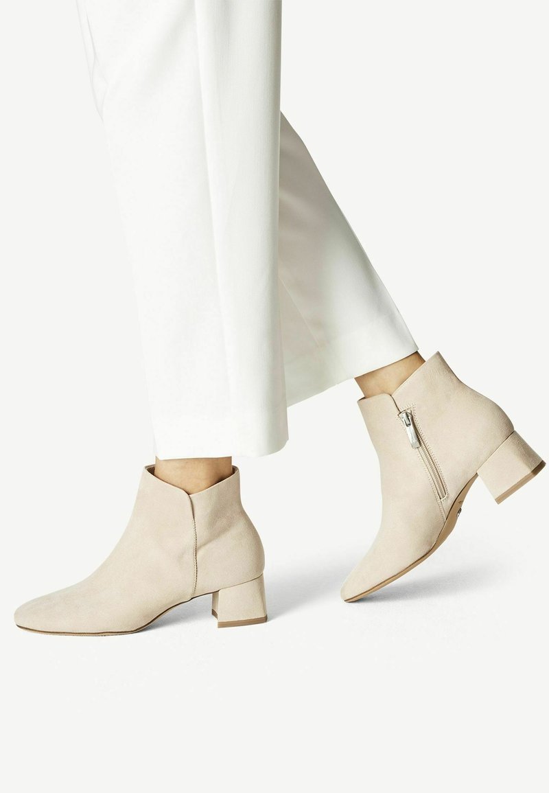 Botines de ante beige con tacón cuadrado, diseño de punta afilada y detalle de cremallera lateral, combinados con pantalones de pierna ancha blancos.