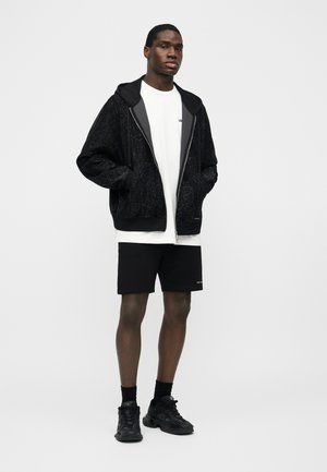 Moški stoji, oblečen v črno kapuco z zadrgo s potiskom, belo majico, črne kratke hlače, črne nogavice in črne športne čevlje, na enobarvnem ozadju.