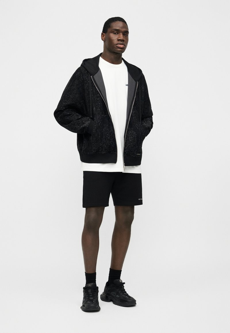 Bărbat în picioare purtând hanorac negru cu fermoar și model, tricou alb, șort negru, șosete negre și încălțăminte sport neagră pe un fundal simplu.