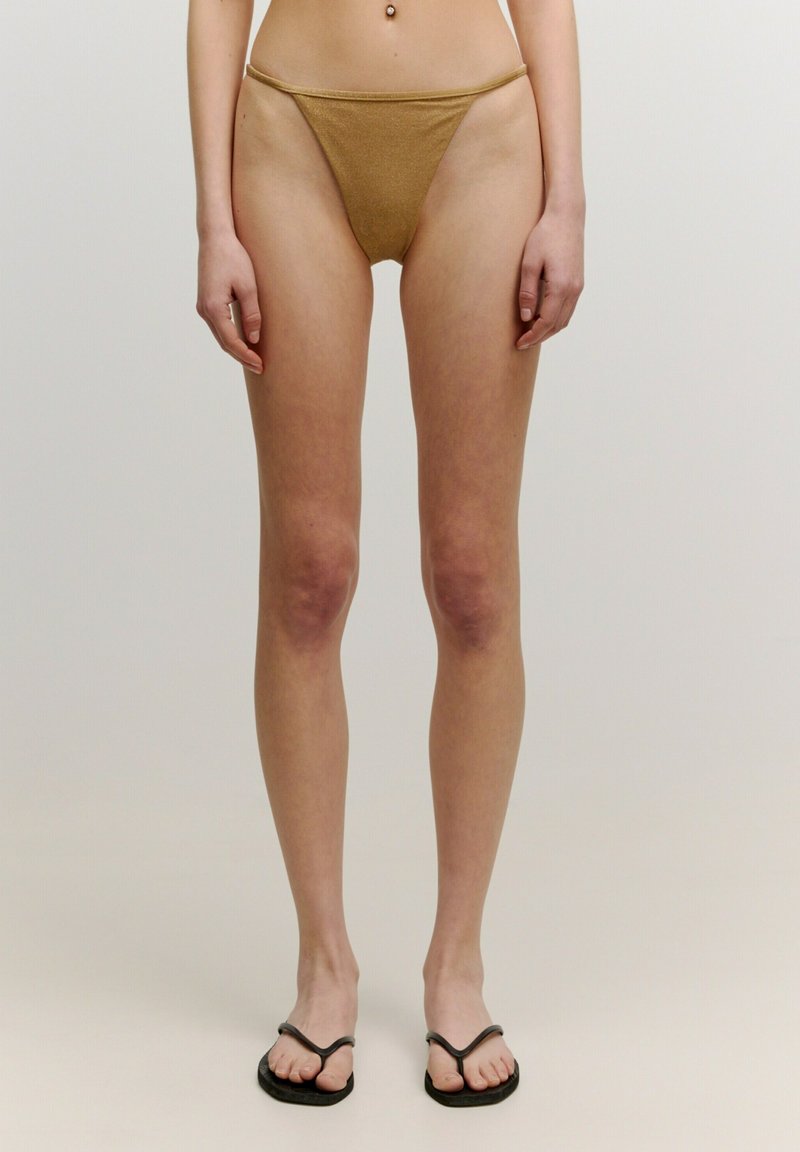 Goldfarbener, metallischer Tanga-Bikiniunterteil mit minimaler Bedeckung, der eine glatte Textur und eine dünne Taille aufweist. Schwarze Flip-Flops mit V-förmigen Riemen.