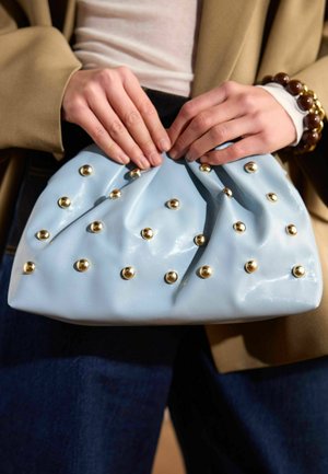 Mani che tengono una pochette azzurro chiaro decorata con borchie dorate, indossata con una giacca beige, una maglietta bianca e pantaloni scuri.