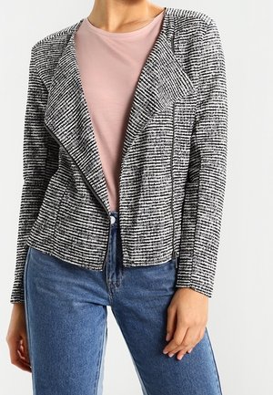 Woman wearing a black and white striped textured blazer over a light pink top and blue jeans, standing against a plain background.