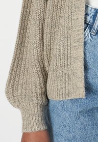 Beige gebreide cardigan met pofmouwen, een gestructureerd patroon en geribbelde manchetten, gecombineerd met lichtblauwe spijkerbroeken.