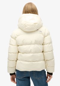 Giacca a vento color crema, con cappuccio e design a righe orizzontali. Il materiale appare liscio con una minima presenza di branding vicino al collo.