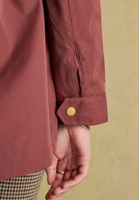 Bordowa kurtka o gładkim wykończeniu, z detalem mankietu na zatrzask i wytłoczonym logo na rękawie. Zbliżenie na teksturowany materiał.