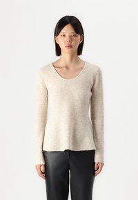 Pull en maille côtelée beige avec un col dégagé, des manches longues et une coupe peplum, réalisé dans un tissu doux et texturé. Associé à un pantalon noir.