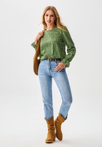 Groene kanten blouse met bloemenpatroon, blauwe spijkerbroek en bruine suède enkellaarsjes. Model draagt een bruine tas en toont lange mouwen met gerimpelde manchetten.