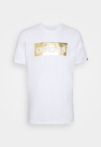 Camiseta de algodón blanca con un logo rectangular metálico dorado que dice "adidas" en blanco, mangas cortas y escote redondo.