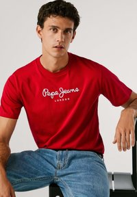 Rdeča bombažna majica s kratkimi rokavi in okroglim izrezom, ki ima bel logotip "Pepe Jeans London", v kombinaciji s svetlo modrimi kavbojkami.