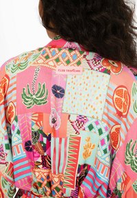 Wielokolorowa koszula w stylu patchwork z wyraźnymi tropikalnymi wzorami, zabawnymi fakturami oraz widoczną metką z napisem "CLUB TROPICANA" na plecach.