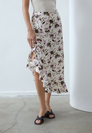 Femme portant une jupe midi blanche à fleurs violettes et noires avec une fente à volants, associée à des sandales à talons ouvertes noires.