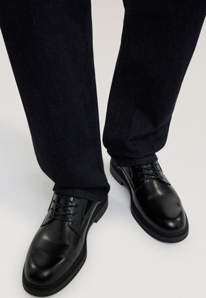 Scarpe nere in pelle lucida con lacci indossate con pantaloni di jeans scuri su uno sfondo neutro.