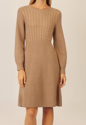 Mujer que lleva un vestido de punto color camello, de manga larga y largo hasta la rodilla, con un patrón de punto trenzado en la parte superior del pecho y punto liso abajo.