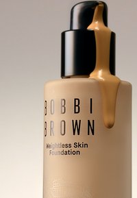 Bobbi Brown Weightless Skin Foundation i en beige flaske med en sort pumpe. Flydende fundament drypper fra dyse, hvilket viser en glat konsistens.