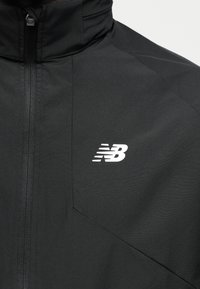 Μαύρο μπουφάν με κάθετο φερμουάρ μπροστά και λευκό λογότυπο New Balance στην αριστερή πλευρά του στήθους, ορατός ψηλός γιακάς.