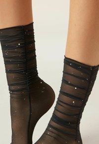 Calze nere trasparenti con arricciature orizzontali, punteggiate da piccoli punti metallici, che creano un aspetto texturizzato e decorativo.