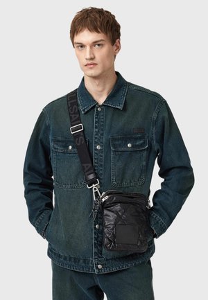Giacca di jeans verde scuro con due tasche frontali e colletto, abbinata a una borsa a tracolla nera trapuntata con una patch logo.