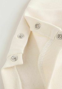 Tela de color crema con broches de presión plateados a lo largo del borde, que presenta una textura acanalada suave y un forro interior liso.