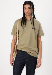 Oliven grønn bomull t-skjorte med korte ermer, med en svart grafisk logo på brystet, kombinert med mørkeblå jeans og en beige crossover veske.