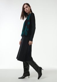 Schwarzes langärmliges Kleid mit fließender Silhouette, kombiniert mit einem türkisfarbenen gemusterten Schal und kniehohen schwarzen Lederstiefeln, die für einen eleganten Look sorgen.