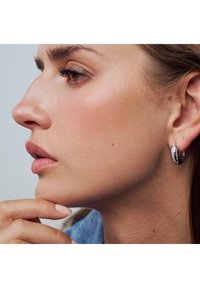 Silverfärgade ringörhängen med ett strukturerat mönster och små strassstenar. Modellens hand rör vid hakan medan hon visar upp örhängena.