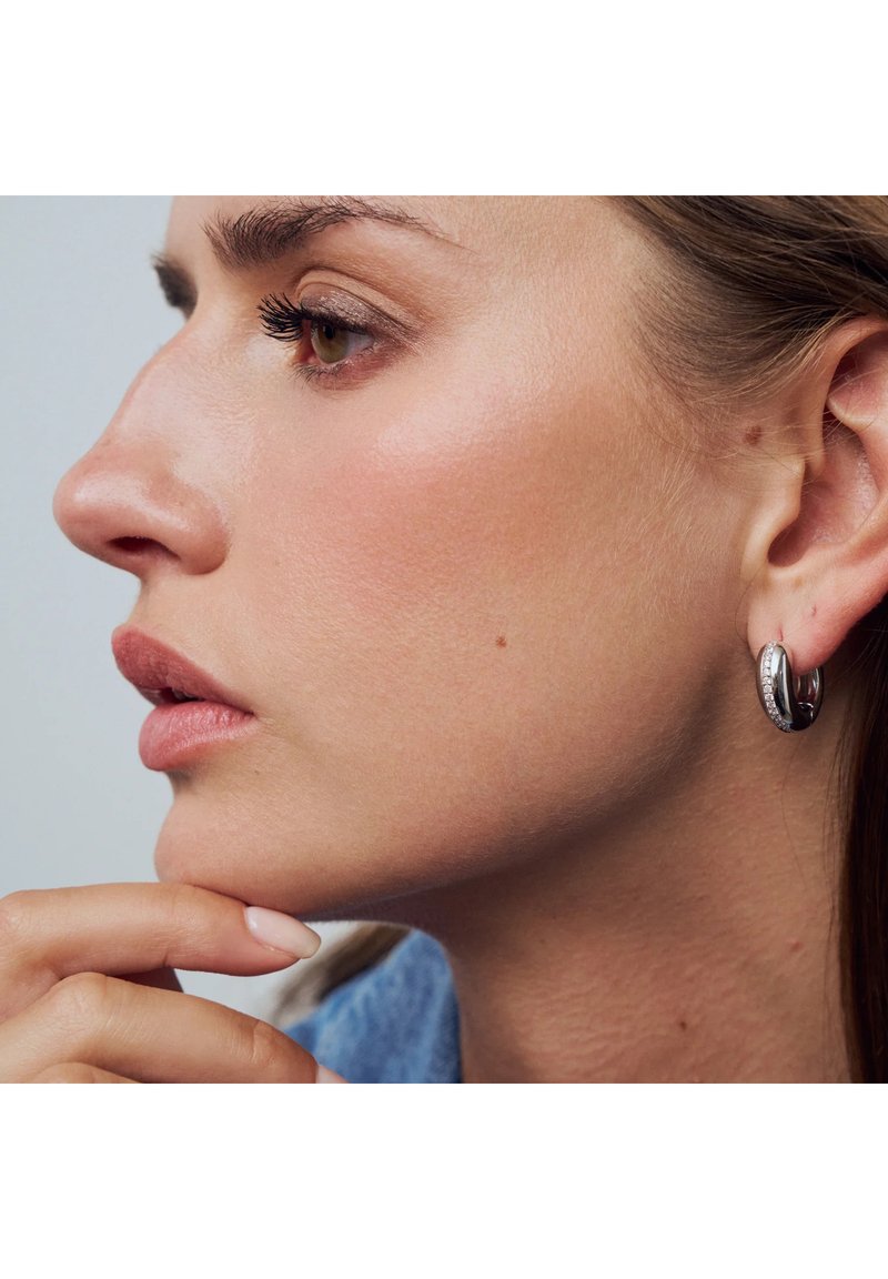 Silverfärgade ringörhängen med ett strukturerat mönster och små strassstenar. Modellens hand rör vid hakan medan hon visar upp örhängena.