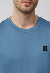 Niebieska bawełniana koszulka z okrągłym dekoltem, z małą czarną metką widoczną po lewej stronie. Gładka tekstura, casualowy design.