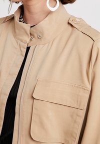 Veste utilitaire beige avec col montant, fermeture éclair devant et grande poche poitrine. Comprend des boutons-pression aux épaules et une texture lisse.