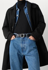Abrigo trinchera negro llevado sobre un suéter de cuello alto oscuro y una chaqueta de mezclilla azul, combinado con jeans azules y un cinturón negro, mostrando un look en capas.