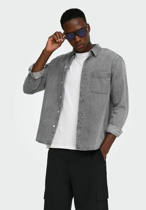 Jeune homme ajustant des lunettes de soleil à verres teintés bleus, portant une chemise grise à boutons sur un t-shirt blanc, avec un pantalon cargo noir sur un fond uni.