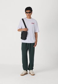 Black crossbody bag, white oversized t-shirt with red text, dark green pants, and cream sneakers with gum soles. Characterized by relaxed fit.