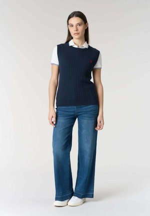 Giovane donna che indossa un maglione senza maniche lavorato a trecce blu navy sopra una polo bianca, jeans larghi a gamba larga blu e sneakers bianche, in piedi contro uno sfondo semplice.