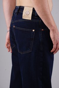 Dunkelblaue Jeans aus strukturiertem Stoff, mit mittelhohem Bund und detaillierten Nähten. Kupferne Knöpfe akzentuieren die Gesäßtaschen.