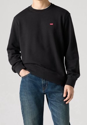 Mężczyzna w gładkiej czarnej bluzie z okrągłym dekoltem i małym czerwonym logo Levi's na klatce piersiowej oraz niebieskich dżinsach, ręka w kieszeni.
