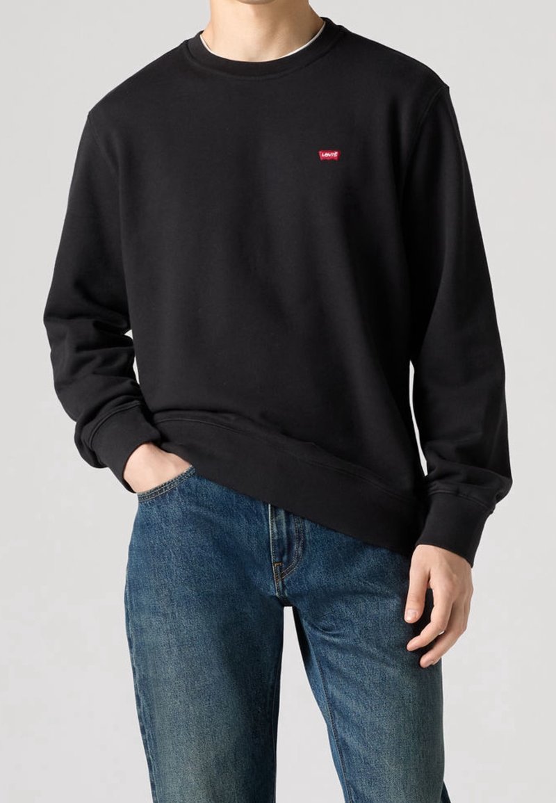 Homme portant un sweat-shirt noir uni à col rond avec un petit logo rouge Levi's sur la poitrine et un jean en denim bleu, main dans la poche.