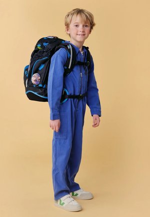 Junge im blauen Overall trägt einen schwarz-blauen Rucksack, weiße Turnschuhe, steht vor einem beigen Hintergrund und lächelt leicht.