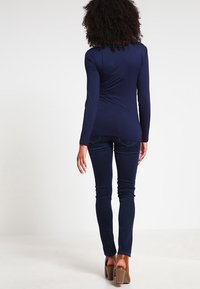 Námořnický svetr s dlouhým rukávem vyrobený z elastické tkaniny, kombinovaný s tmavými skinny džíny. Model stojí zády k divákovi, čímž zdůrazňuje střih a tvar.